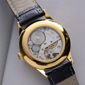 K&ouml;nnte beinhalten: Nahaufnahme einer goldfarbenen Armbanduhr mit transparentem Geh&auml;useboden, der das komplizierte mechanische Uhrwerk zeigt. Die Uhr hat ein schwarzes Lederarmband. Das Zifferblatt zeigt den Text "2609.HA" und "NS".