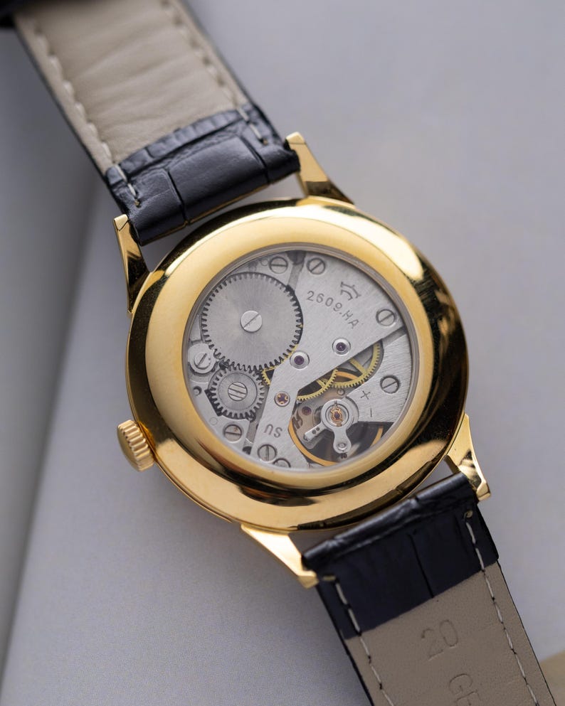 K&ouml;nnte beinhalten: Nahaufnahme einer goldfarbenen Armbanduhr mit transparentem Geh&auml;useboden, der das komplizierte mechanische Uhrwerk zeigt. Die Uhr hat ein schwarzes Lederarmband. Das Zifferblatt zeigt den Text "2609.HA" und "NS".