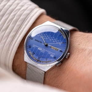 Peut inclure: Une montre Raketa argentée avec un cadran bleu et un bracelet en maille. Le cadran affiche la date et le mois, avec le texte "RAKETA" en haut. La montre est portée au poignet.