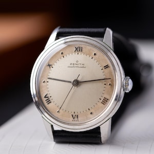 May include: A vintage Zenith automatic wristwatch with a silver-tone case and a cream-colored face. The watch features black Roman numerals, black hands, and a black leather band. The watch face has the text "ZENITH automatic" and "SWISS".