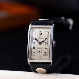 Peut inclure: Une montre Omega vintage avec un boîtier rectangulaire argenté et un bracelet en cuir noir. Le cadran est blanc cassé avec des chiffres et des aiguilles noirs. La montre a un petit cadran de secondes et le logo Omega.