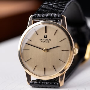 May include: A gold-toned Universal Genève wristwatch with a black leather strap. The watch face is a light gold colour with black stick hour markers and hands. The watch has the text "UNIVERSAL GENEVE" on the face. The strap is textured.