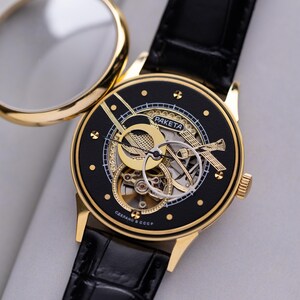 K&ouml;nnte beinhalten: Eine goldfarbene Armbanduhr mit schwarzem Zifferblatt und schwarzem Lederarmband. Das Zifferblatt zeigt komplizierte goldfarbene Zahnr&auml;der und das Wort "RAKETA". Die Uhr ist ge&ouml;ffnet und zeigt eine Lupe.