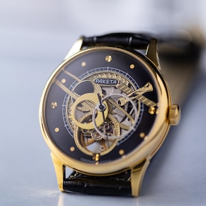 K&ouml;nnte beinhalten: Eine goldfarbene Armbanduhr mit schwarzem Zifferblatt und sichtbaren Innenmechanismen. Die Uhr tr&auml;gt den Markennamen "PAKETA" und ist an einem schwarzen Lederarmband befestigt. Die Uhr hat ein klassisches, elegantes Design.