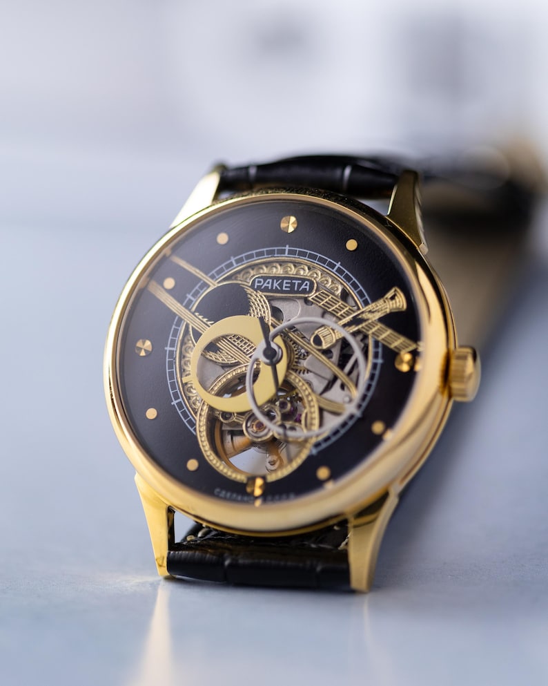 K&ouml;nnte beinhalten: Eine goldfarbene Armbanduhr mit schwarzem Zifferblatt und sichtbaren Innenmechanismen. Die Uhr tr&auml;gt den Markennamen "PAKETA" und ist an einem schwarzen Lederarmband befestigt. Die Uhr hat ein klassisches, elegantes Design.
