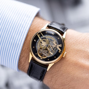 K&ouml;nnte beinhalten: Eine goldfarbene Armbanduhr mit schwarzem Zifferblatt und sichtbarem Uhrwerk. Das Armband ist aus schwarzem Leder. Das Wort "Raketa" ist auf dem Zifferblatt zu sehen. Die Uhr befindet sich an einem Handgelenk.