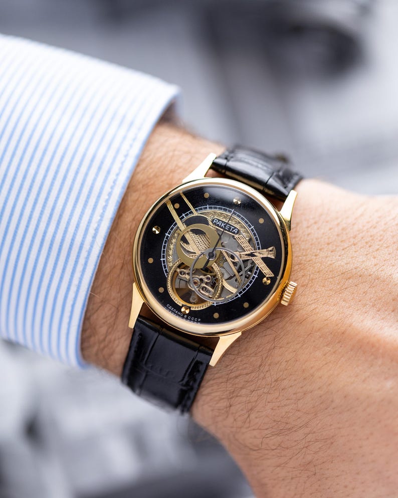 K&ouml;nnte beinhalten: Eine goldfarbene Armbanduhr mit schwarzem Zifferblatt und sichtbarem Uhrwerk. Das Armband ist aus schwarzem Leder. Das Wort "Raketa" ist auf dem Zifferblatt zu sehen. Die Uhr befindet sich an einem Handgelenk.