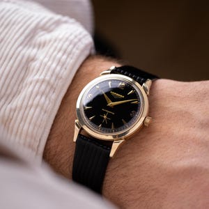 Peut inclure: Une montre-bracelet dorée avec un cadran noir et un bracelet noir. Le cadran de la montre présente des aiguilles et des marqueurs dorés, avec la marque "Longines" visible. La montre est portée au poignet, avec une manche de chemise blanche en arrière-plan.