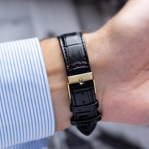 K&ouml;nnte beinhalten: Nahaufnahme eines schwarzen Leder-Uhrenarmbands mit einer goldfarbenen Schnalle. Das Uhrenarmband ist an einem Handgelenk befestigt, und die Person tr&auml;gt ein blau-wei&szlig; gestreiftes Hemd.