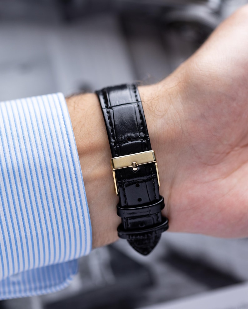 K&ouml;nnte beinhalten: Nahaufnahme eines schwarzen Leder-Uhrenarmbands mit einer goldfarbenen Schnalle. Das Uhrenarmband ist an einem Handgelenk befestigt, und die Person tr&auml;gt ein blau-wei&szlig; gestreiftes Hemd.