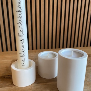 Könnte beinhalten: Drei weiße Kerzen, darunter eine mit dem schwarzen Schriftzug "ein kleines Dankeschön", stehen auf einer Holzoberfläche. Der Hintergrund besteht aus Holzlatten.