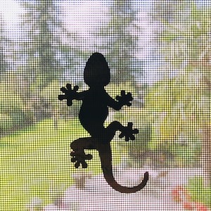 Pode incluir: Uma silhueta preta de um gecko está presa a uma tela, com uma cena externa desfocada visível através da malha. O gecko está em uma pose de escalada, com a cauda enrolada. O fundo apresenta folhagem verde e um toque de céu azul.