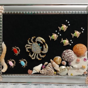 May include: A rectangular silver-toned frame containing a black velvet background decorated with various sea-themed embellishments, including a large rhinestone-encrusted crab, smaller colorful crab pins, fish figures, and an assortment of seashells.