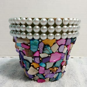 May include: A decorative flower pot with a mosaic design. The pot is adorned with colorful, irregularly shaped pieces of shell in shades of pink, blue, purple, and gold. The rim is embellished with multiple rows of white pearls.