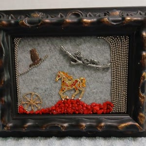 May include: A decorative framed wall art piece featuring a gray felt background with a gold-toned horse figurine, a silver-toned bird branch, a small owl, a gold wheel, and a pile of red stones, all enclosed in a dark brown ornate frame.