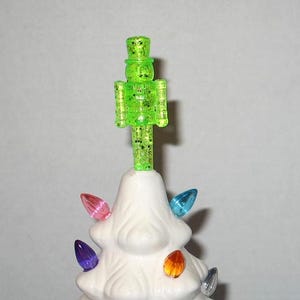 May include: A white ceramic Christmas tree with colorful light bulbs and a green glitter nutcracker topper.