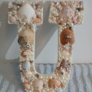 May include: A decorative letter "U" made of white material, adorned with various seashells and faux pearls. The seashells are in shades of white, tan, and brown. The pearls are small and clustered around the edges and within the letter's form.