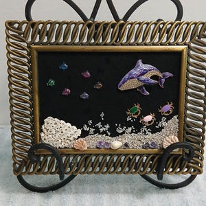 May include: A rectangular framed wall art piece featuring a black velvet background with a purple dolphin brooch, small colorful fish, and three crab brooches. The bottom includes white coral, sand, and small seashells, all encased in a twisted bronze frame.