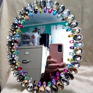 May include: A round mirror framed by a border of multi-colored, faceted, oval-shaped glass gems in shades of clear, amber, blue, pink, and purple. The reflective surface shows a reflection of a pink smartphone and a beige filing cabinet.