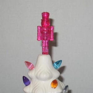 May include: A white ceramic Christmas tree with a pink glitter nutcracker topper and colorful light bulbs.