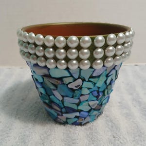 May include: A decorative terracotta flower pot. The top rim is adorned with three rows of white faux pearls. The lower portion is covered in a mosaic of blue and turquoise shell pieces. The interior is a natural terracotta color.