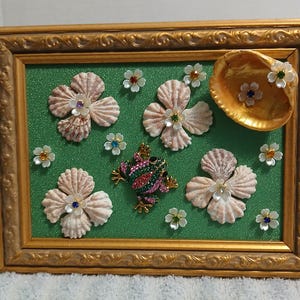 May include: A rectangular gold-toned frame containing a green textured background decorated with four shell flowers, several small white floral accents with colored centers, a gold shell, and a central frog brooch with pink and green crystals.