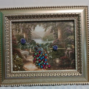 May include: A rectangular gold-toned frame with a beaded border containing a landscape print of a garden terrace overlooking a lake. A jeweled peacock brooch with red, blue, and green rhinestones is attached to the center of the print.