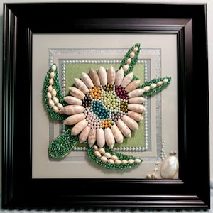 May include: Framed artwork featuring a sea turtle design. The turtle's shell is made of various seashells and colorful beads. The frame is black, and the background is light green and gray.
