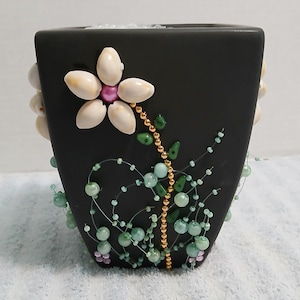 May include: Black square ceramic vase decorated with a flower made of seashells and a beaded stem. The vase is adorned with green and blue beads, and a gold beaded stem. The flower has a pink center.