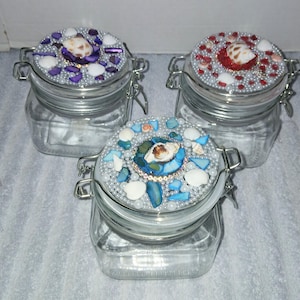 May include: Three glass jars with decorative lids. Each lid is adorned with seashells, beads, and other embellishments in shades of purple, red, and blue. The jars have metal clasps.