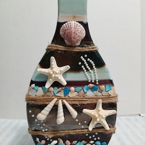 May include: A decorative vase with a coastal theme. The vase is adorned with seashells, starfish, and small stones in shades of blue, white, and brown. Jute rope accents the design, and a large seashell is affixed near the top.