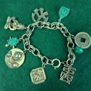May include: A gold-tone metal charm bracelet featuring various dangling charms, including a round coin with a square hole, a rectangular charm with Asian characters, a circular medallion with a relief figure, and several green translucent beads.