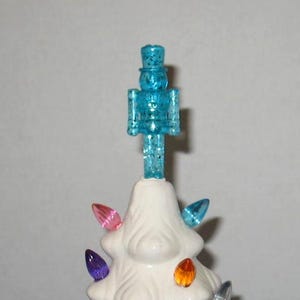 May include: A white ceramic Christmas tree with colorful light bulbs and a blue glitter nutcracker on top.