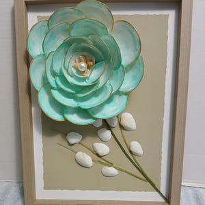 May include: A framed wall art piece featuring a large, three-dimensional flower crafted from light turquoise shells with gold-painted edges. The center contains small shells and a white pearl. White shells and green stems are arranged below.