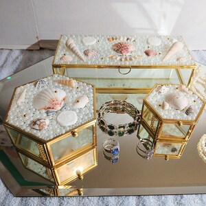May include: Three glass jewelry boxes with gold trim, decorated with seashells and faux pearls. A rectangular box and two hexagonal boxes are displayed with jewelry, including rings and a bracelet, on a mirrored surface.