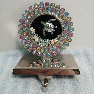May include: Silver-toned stocking holder with a circular design featuring a sea turtle on a black background, surrounded by iridescent jewels. The base is rectangular with additional jewels.