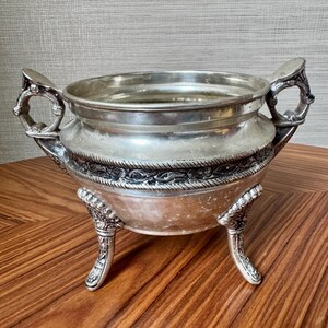May include: A silver-toned metal bowl featuring two ornate handles on opposite sides and three decorative legs. The bowl has a wide, circular opening and a band of intricate, raised floral and rope-like detailing encircling the midsection.