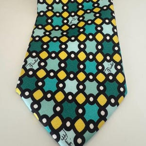May include: A necktie featuring a geometric pattern of interlocking black circles and yellow diamonds on a background of teal and light blue squares. The word "Fiulio" is printed in a cursive black script throughout the fabric design.