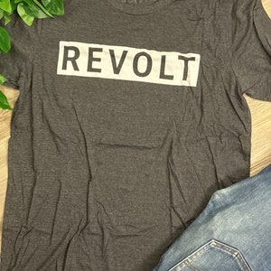 May include: A dark gray t-shirt with a white rectangle and the word "REVOLT" printed in white.