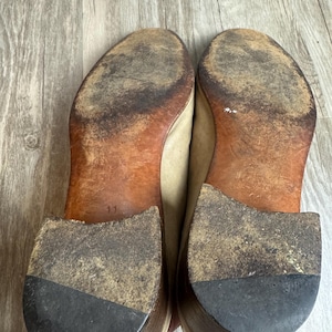 May include: A pair of worn beige leather shoes, soles facing up. The soles are brown with significant wear and dirt, and the heels are dark gray. The number "11" is visible on one sole. The shoes appear to be well-used.