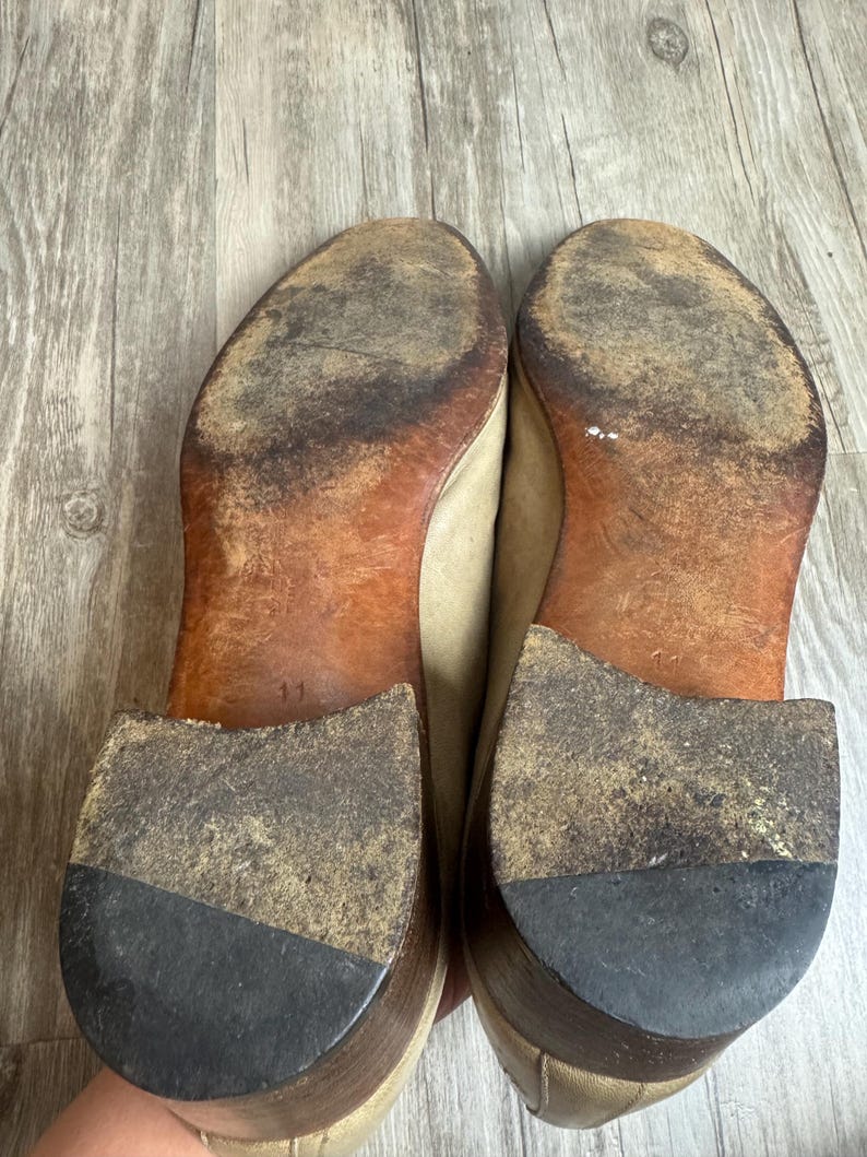 May include: A pair of worn beige leather shoes, soles facing up. The soles are brown with significant wear and dirt, and the heels are dark gray. The number "11" is visible on one sole. The shoes appear to be well-used.