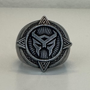 May include: A silver-colored ring featuring a detailed design of a stylized panther head within a circular emblem. The ring has a textured band and geometric accents, with a triangular element at the top.