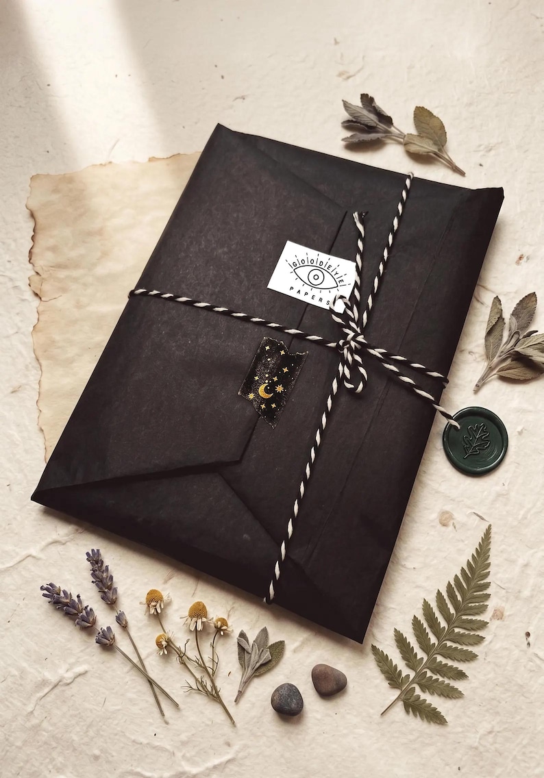May include: A rectangular package wrapped in black paper, tied with black and white baker's twine. A white sticker with an eye illustration and the text "PAPERS" is attached. A dark green wax seal with a leaf imprint hangs from the twine.