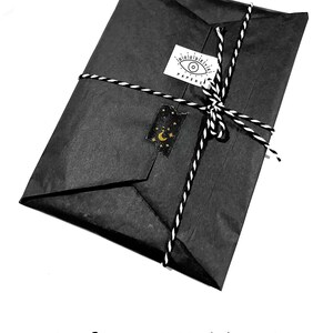 May include: A black parcel wrapped in tissue paper and tied with black and white twine. A sticker with an eye design and the word "PAPERS" is on the package. A gold and black sticker with a moon and stars is also on the package.