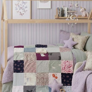 Può includere: Una trapunta patchwork in un ambiente di asilo nido. La trapunta presenta quadrati di vari tessuti nei toni del viola, grigio e rosa. Sono visibili anche un cuscino a forma di stella e un letto con struttura in legno. Una giostrina con stelle e lune è appesa sopra il letto.