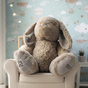 Puede incluir: Un gran conejo de peluche marrón, con orejas largas y detalles de pana, sentado en un sillón crema. Las orejas y patas del conejo tienen detalles bordados. El fondo presenta un papel pintado azul claro con globos aerostáticos y nubes.