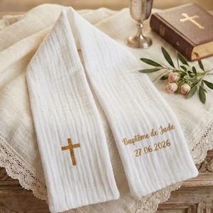 Peut inclure: Étole de baptême blanche avec une croix dorée et le texte "Baptême de Jade 27.06.2026". L'étole est drapée sur un tissu bordé de dentelle, avec un calice argenté et un livre avec une croix en arrière-plan.