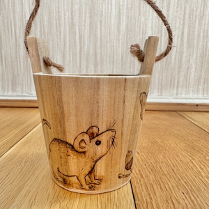 May include: A small, wooden bucket with a rope handle. The bucket features a detailed wood-burned illustration of a mouse and an acorn. The bucket is made of light-colored wood and is suitable for decorative use.