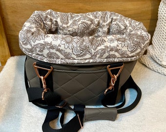 May include: A gray quilted pet carrier with a white floral patterned lining. The carrier has two adjustable straps and a top handle.