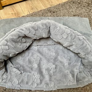 May include: A gray pet bed with a soft, plush lining featuring a paw print design. The bed is made of a corduroy fabric.